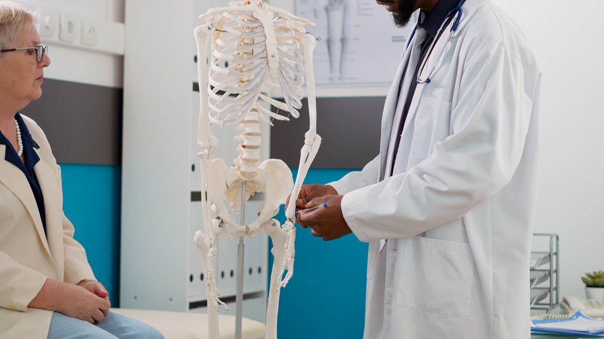 Physician examining human skeleton in cabinet at checkup visit, explaining anatomy bones diagnosis to elderly patient. Specialist analyzing spinal cord to help with orthopedic treatment.